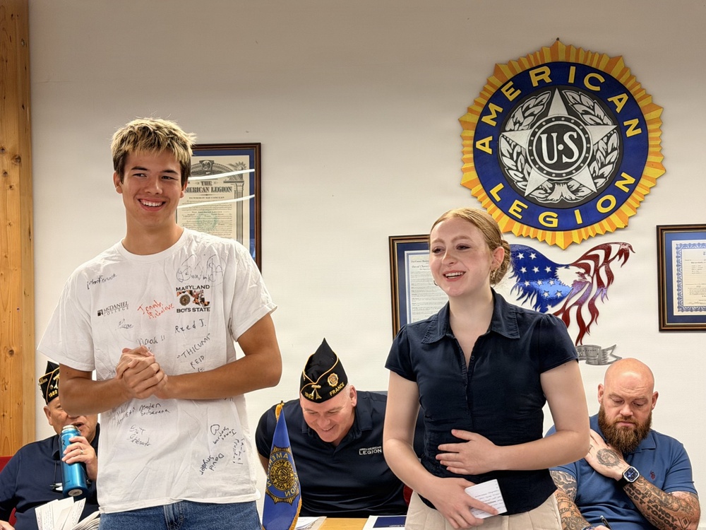 Two young people stand smiling at what appears to be an American Legion post meeting or recognition event. The young man is wearing a white t-shirt covered in signatures, labeled 'Maryland Boys State,' suggesting he recently attended a Boys State program. They appear to be receiving recognition or presenting to the group gathered at the post.