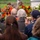 Attendees at the LT John S Thompson Bridge Ceremony, Operation Market Garden Commemorations 2026 A memorial or commemoration ceremony is taking place outdoors, with an audience of mostly elderly attendees seated and facing a monument or memorial marker. Colorful floral wreaths in orange, red, and white are prominently displayed near the monument. One attendee wearing a navy blue cap and an American flag pin is capturing the ceremony on a smartphone.