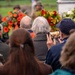 A memorial or commemoration ceremony is taking place outdoors, with an audience of mostly elderly attendees seated and facing a monument or memorial marker. Colorful floral wreaths in orange, red, and white are prominently displayed near the monument. One attendee wearing a navy blue cap and an American flag pin is capturing the ceremony on a smartphone.