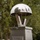 A Memorial is Displayed at the LT John S Thompson Bridge Ceremony, Operation Market Garden Commemorations 2026 A polished stainless steel memorial sculpture shaped like a parachute sits atop a pedestal monument. The base bears a dedication plaque reading 'John S. Thompson-Brug' with a date referencing 1944, suggesting a World War II commemoration. The monument is situated outdoors, surrounded by trees, with flags reflected in the dome's mirrored surface.