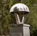 A polished stainless steel memorial sculpture shaped like a parachute sits atop a pedestal monument. The base bears a dedication plaque reading 'John S. Thompson-Brug' with a date referencing 1944, suggesting a World War II commemoration. The monument is situated outdoors, surrounded by trees, with flags reflected in the dome's mirrored surface.