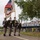 21st Theater Sustainment Command Color Guard Presents the Colors at the LT John S Thompson Bridge Ceremony, Operation Market Garden Commemorations 2026 A U.S. Army color guard unit marches in formation along a paved path, carrying the American flag and a United States Army unit guidon. The ceremony appears to take place near a bridge in what looks like a European setting, suggested by the Dutch-style cycling infrastructure signs and bicycle parking visible on the right. The soldiers are dressed in Army Service Uniforms (ASU) in olive/tan tones with white gloves.
