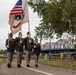 A U.S. Army color guard unit marches in formation along a paved path, carrying the American flag and a United States Army unit guidon. The ceremony appears to take place near a bridge in what looks like a European setting, suggested by the Dutch-style cycling infrastructure signs and bicycle parking visible on the right. The soldiers are dressed in Army Service Uniforms (ASU) in olive/tan tones with white gloves.