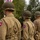 WWII Reenactors Stand at Attention at the at the LT John S Thompson Bridge Ceremony, Operation Market Garden Commemorations 2026 The image shows multiple individuals wearing WWII-era U.S. Army uniforms with M1 steel helmets and 82nd Airborne Division ('AA') shoulder patches. They appear to be standing in formation at what looks like a commemorative event or reenactment. Flags and civilian spectators are visible in the background.