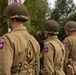 The image shows multiple individuals wearing WWII-era U.S. Army uniforms with M1 steel helmets and 82nd Airborne Division ('AA') shoulder patches. They appear to be standing in formation at what looks like a commemorative event or reenactment. Flags and civilian spectators are visible in the background.