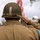 WWII Reenactor Salutes at the LT John S Thompson Bridge Ceremony, Operation Market Garden Commemorations 2026 A soldier or reenactor wearing WWII-era U.S. Army uniform is photographed from behind while rendering a salute. The subject wears an M1 steel helmet with camouflage netting and an olive drab field jacket bearing a U.S. flag patch on the right sleeve. An American flag is visible in the blurred background, suggesting a commemorative or ceremonial event.