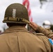 A soldier or reenactor wearing WWII-era U.S. Army uniform is photographed from behind while rendering a salute. The subject wears an M1 steel helmet with camouflage netting and an olive drab field jacket bearing a U.S. flag patch on the right sleeve. An American flag is visible in the blurred background, suggesting a commemorative or ceremonial event.
