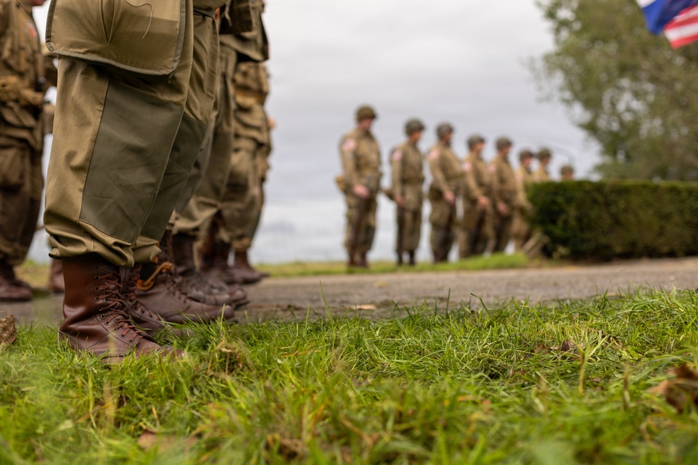 The image shows a group of individuals dressed in WWII-era U.S. Army uniforms standing in formation on a grassy area. The foreground features a close-up ground-level perspective of brown leather combat boots and olive drab trousers, while a line of similarly uniformed personnel is visible in the blurred background. An American flag is partially visible in the upper right corner, suggesting a commemorative or reenactment event.