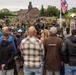 A World War II commemoration ceremony is taking place outdoors, likely in the Netherlands, featuring reenactors in period American military uniforms, vintage military vehicles, and a brass band performing for a large civilian audience. Flags including the Union Jack (United Kingdom) and what appears to be the Dutch flag are prominently displayed on flagpoles. The event appears to be a liberation anniversary memorial gathering, consistent with annual commemorations held in the Netherlands.