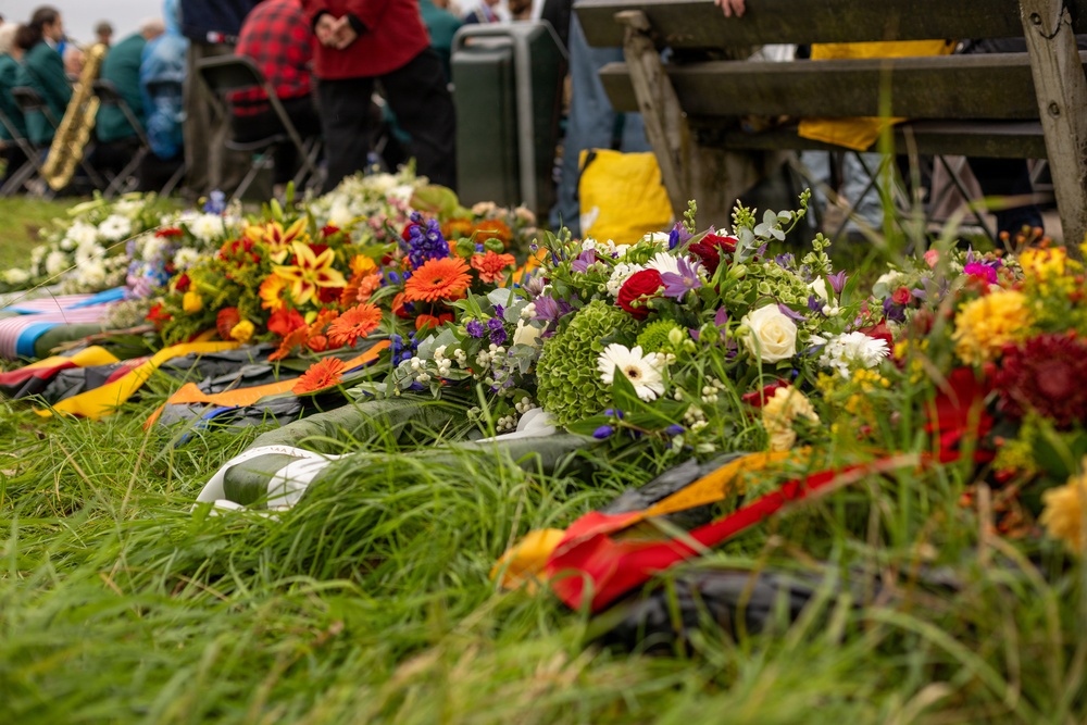 Multiple colorful floral wreaths and bouquets are laid on grass, suggesting a commemorative or memorial ceremony. The arrangements feature a variety of flowers including gerberas, roses, lilies, and hydrangeas with decorative ribbons in red, yellow, and orange. In the blurred background, people are gathered and a saxophone is visible, indicating a formal outdoor event with musical accompaniment.