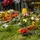 Wreaths are Laid Out at the LT John S Thompson Bridge Ceremony, Operation Market Garden Commemorations 2026 Multiple colorful floral wreaths and bouquets are laid on grass, suggesting a commemorative or memorial ceremony. The arrangements feature a variety of flowers including gerberas, roses, lilies, and hydrangeas with decorative ribbons in red, yellow, and orange. In the blurred background, people are gathered and a saxophone is visible, indicating a formal outdoor event with musical accompaniment.
