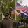 An Attendee Salutes at the LT John S Thompson Bridge Ceremony, Operation Market Garden Commemorations 2026 A person wearing a dark green beret and olive jacket is saluting toward a row of national flags at what appears to be a commemorative or military ceremony. Multiple flags are visible including the United States flag (twice), the United Kingdom Union Jack, and the Netherlands tricolor. A crowd of attendees is visible in the background.