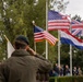 A person wearing a dark green beret and olive jacket is saluting toward a row of national flags at what appears to be a commemorative or military ceremony. Multiple flags are visible including the United States flag (twice), the United Kingdom Union Jack, and the Netherlands tricolor. A crowd of attendees is visible in the background.