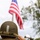 WWII Reenactor Salutes at the LT John S Thompson Bridge Ceremony, Operation Market Garden Commemorations 2026 A soldier or reenactor wearing WWII-era U.S. Army uniform and M1 helmet with camouflage netting is saluting an American flag. The subject is photographed from behind, with an American flag patch visible on the left sleeve of the olive drab jacket. The background shows a blurred American flag on a pole, trees, and other people gathered at what appears to be a commemorative or ceremonial event.