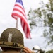 A soldier or reenactor wearing WWII-era U.S. Army uniform and M1 helmet with camouflage netting is saluting an American flag. The subject is photographed from behind, with an American flag patch visible on the left sleeve of the olive drab jacket. The background shows a blurred American flag on a pole, trees, and other people gathered at what appears to be a commemorative or ceremonial event.