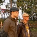 Two men stand outdoors in what appears to be a field or event area cordoned off with red-and-white barrier tape. The younger man on the left wears a military-style black beret, olive jacket, and camouflage trousers, while the older man on the right wears a tan baseball cap with a '101st Airborne' screaming eagle patch, a leather jacket, and a hoodie with military-themed graphics. Both are looking in the same direction, suggesting they are observing something off-camera.