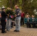 A commemorative ceremony is taking place outdoors, likely a memorial or liberation remembrance event. A scout wearing a badge-covered uniform is presenting a large floral wreath adorned with small flags to an official in a dark uniform and white cap. A band in green jackets is seated to the right, and an audience of attendees watches the proceedings.