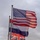 Flags are Flown at the LT John S Thompson Bridge Ceremony, Operation Market Garden Commemorations 2026 Three national flags are flying from a single tall flagpole against an overcast sky. The flags are actively waving in the wind, with the United States flag at the top, the French Tricolor in the middle, and the United Kingdom Union Jack at the bottom. A small tree is partially visible in the lower left corner.