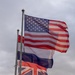 Three national flags are flying from a single tall flagpole against an overcast sky. The flags are actively waving in the wind, with the United States flag at the top, the French Tricolor in the middle, and the United Kingdom Union Jack at the bottom. A small tree is partially visible in the lower left corner.