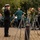 U.S. Navy Attendee Renders Honors at the LT John S Thompson Bridge Ceremony, Operation Market Garden Commemorations 2026 A military memorial ceremony is taking place outdoors, featuring a naval officer in dark dress uniform rendering a salute toward a floral wreath on a stand. A civilian brass band in green jackets provides musical accompaniment in the background, while a second military officer stands at attention near a monument. The wreath displays red, white, and blue flowers along with what appears to be an American flag ribbon.