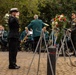 A military memorial ceremony is taking place outdoors, featuring a naval officer in dark dress uniform rendering a salute toward a floral wreath on a stand. A civilian brass band in green jackets provides musical accompaniment in the background, while a second military officer stands at attention near a monument. The wreath displays red, white, and blue flowers along with what appears to be an American flag ribbon.
