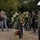 Attendees Lay a Wreath at the LT John S Thompson Bridge Ceremony, Operation Market Garden Commemorations 2026 A formal memorial ceremony is taking place at an outdoor monument, featuring wreath-laying by civilians and a uniformed military officer reading from a document. A band dressed in green uniforms performs in the background. Two young women carry a floral bouquet toward the memorial, which features a distinctive silver dome-topped pillar with a commemorative plaque.