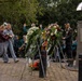 A formal memorial ceremony is taking place at an outdoor monument, featuring wreath-laying by civilians and a uniformed military officer reading from a document. A band dressed in green uniforms performs in the background. Two young women carry a floral bouquet toward the memorial, which features a distinctive silver dome-topped pillar with a commemorative plaque.