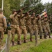A group of re-enactors dressed in WWII-era U.S. Army uniforms are performing a ceremonial rifle salute (volley fire) at what appears to be a commemorative event. The soldiers are lined up in formation, firing rifles skyward, while spectators and photographers observe from the background. A U.S. flag patch and period-accurate equipment suggest this is a memorial or historical re-enactment ceremony.