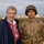 Chloe Gavin and Gavin Ryan Keereweer at the LT John S Thompson Bridge Ceremony, Operation Market Garden Commemorations 2026 Two individuals pose together outdoors at dusk or dawn against a grassy field with bokeh lights in the background. One is an older woman wearing a navy jacket and a red, white, and blue patriotic scarf. The other is a young man dressed in a WWII-era U.S. Army uniform, including an M1 helmet with netting, khaki field jacket, and an 82nd Airborne Division patch on his left shoulder.