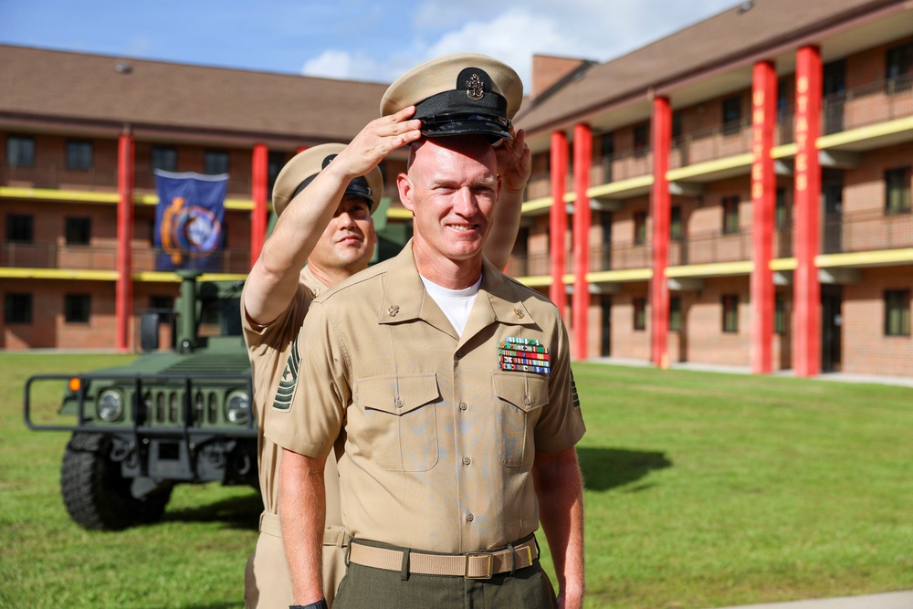 Two U.S. Marine Corps personnel in tan service uniforms are photographed outdoors on what appears to be a military base. One Marine is placing or adjusting a dress cover (cap) on the other Marine's head, suggesting a promotion or ceremonial moment. The setting includes a multi-story brick barracks building with red columns and a military vehicle (HMMWV/Humvee) in the background.