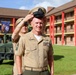 Two U.S. Marine Corps personnel in tan service uniforms are photographed outdoors on what appears to be a military base. One Marine is placing or adjusting a dress cover (cap) on the other Marine's head, suggesting a promotion or ceremonial moment. The setting includes a multi-story brick barracks building with red columns and a military vehicle (HMMWV/Humvee) in the background.