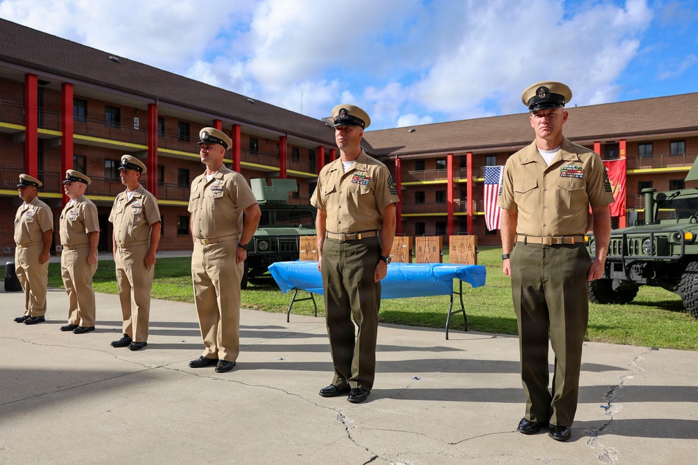 A group of military personnel in tan/khaki service uniforms are standing at attention in formation on a paved courtyard area. The setting appears to be a formal military ceremony or parade at a base installation, with an American flag and what appears to be a Marine Corps flag visible in the background. Military vehicles, including Humvees, are parked nearby.