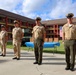 A group of military personnel in tan/khaki service uniforms are standing at attention in formation on a paved courtyard area. The setting appears to be a formal military ceremony or parade at a base installation, with an American flag and what appears to be a Marine Corps flag visible in the background. Military vehicles, including Humvees, are parked nearby.