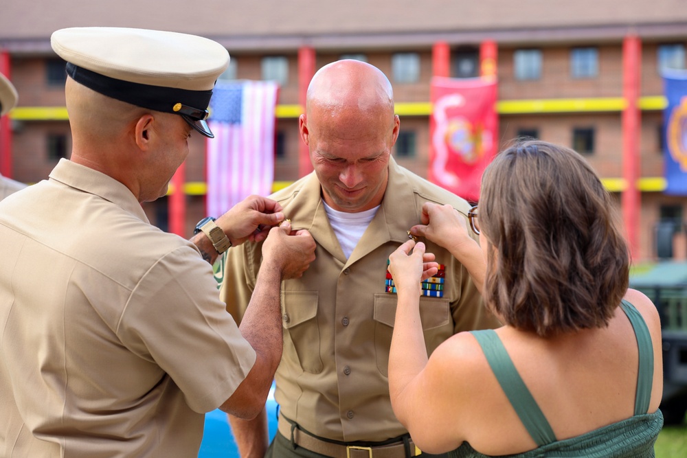 A military promotion or award ceremony is taking place outdoors at what appears to be a U.S. Marine Corps installation. Two individuals — a uniformed military officer and a civilian woman — are simultaneously pinning insignia or rank devices onto the collar of a bald servicemember in a tan uniform. The setting includes visible U.S. and Marine Corps flags in the background.