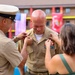 A military promotion or award ceremony is taking place outdoors at what appears to be a U.S. Marine Corps installation. Two individuals — a uniformed military officer and a civilian woman — are simultaneously pinning insignia or rank devices onto the collar of a bald servicemember in a tan uniform. The setting includes visible U.S. and Marine Corps flags in the background.