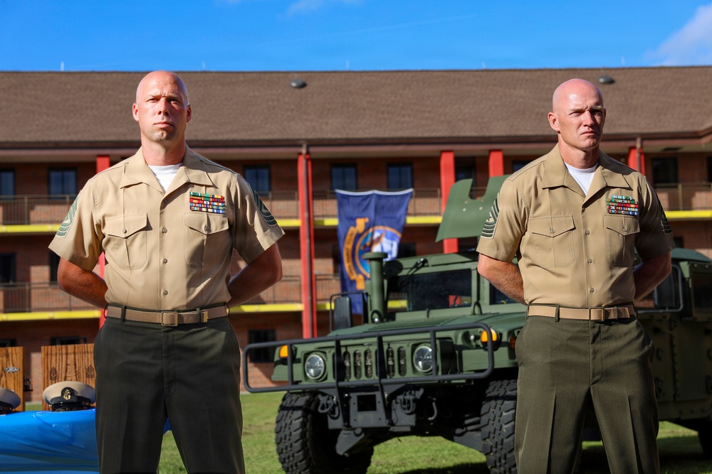 Two bald male military personnel in tan/khaki United States Marine Corps service uniforms stand at parade rest outdoors. Both wear multiple service ribbons and sergeant major or senior NCO rank insignia on their collars. A military HMMWV (Humvee) and a unit guidon/flag are visible in the background behind a multi-story brick building.