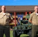 Two bald male military personnel in tan/khaki United States Marine Corps service uniforms stand at parade rest outdoors. Both wear multiple service ribbons and sergeant major or senior NCO rank insignia on their collars. A military HMMWV (Humvee) and a unit guidon/flag are visible in the background behind a multi-story brick building.