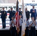 A formal military or civic ceremony is taking place, likely in New York City based on the visible signage referencing 'City of New York.' A youth color guard in light blue uniforms presents the American flag and additional flags while armed with ceremonial rifles, as uniformed Coast Guard officers and civilians on a raised stage render salutes and hand-over-heart gestures. The setting appears to be near a waterfront, with a large vessel visible in the background.