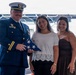 A uniformed military or Coast Guard officer is presenting a folded American flag to two young women during what appears to be a formal ceremony. The officer is wearing a dress blue uniform with multiple ribbons, medals, and gold sleeve stripes indicating officer rank. The setting appears to be outdoors near a vessel or dock structure.