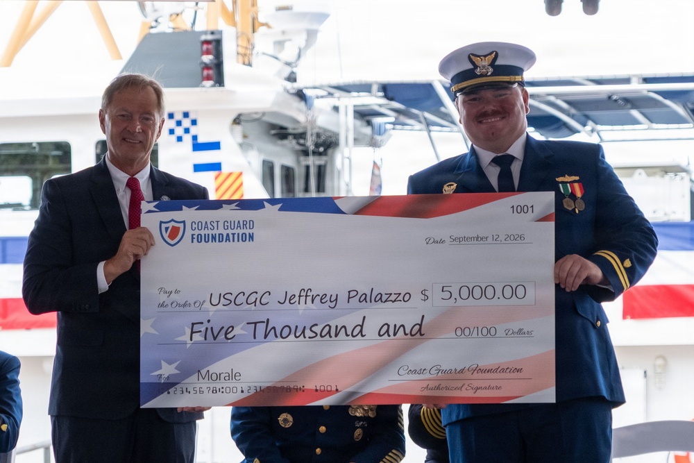 Two individuals are holding a large ceremonial check from the Coast Guard Foundation made out to USCGC Jeffrey Palazzo for $5,000.00, dated September 12, 2026. The check is designated for 'Morale' purposes. The event appears to be a formal military commissioning or dedication ceremony taking place near a Coast Guard vessel.