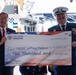 Two individuals are holding a large ceremonial check from the Coast Guard Foundation made out to USCGC Jeffrey Palazzo for $5,000.00, dated September 12, 2026. The check is designated for 'Morale' purposes. The event appears to be a formal military commissioning or dedication ceremony taking place near a Coast Guard vessel.
