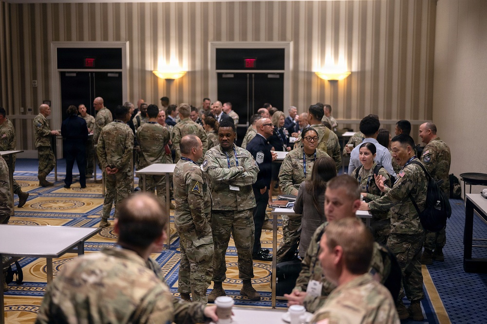 A large group of military personnel in camouflage uniforms are gathered in what appears to be a hotel ballroom or conference facility. The gathering appears to be a military conference or networking event, with personnel mingling, conversing in small groups, and some holding beverages. A small number of civilians in business attire are also present among the uniformed attendees.