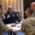 Space Force Connection Time at AFA 2026 A group of military personnel are engaged in a discussion around a standing table in what appears to be a conference or meeting room. One individual wearing a dark blue Air Force dress uniform is gesturing and speaking to others dressed in OCP (Operational Camouflage Pattern) uniforms. A military cap with the name 'JOSLIN' is visible on the table.