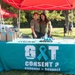 Two individuals stand smiling behind a teal-covered table at an outdoor awareness booth under a red canopy tent. The table displays a 'Got Consent? Consent = Respect' banner featuring a teal awareness ribbon, along with informational materials, giveaway items, and hand sanitizer. The event appears to be a sexual assault awareness or prevention outreach activity held in a grassy outdoor setting.