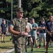A U.S. Marine in full MARPAT camouflage uniform and cover (cap) addresses a group of civilians and possibly military personnel in civilian attire on an outdoor athletic field. The Marine appears to be in a leadership role, speaking to the assembled group who are standing and listening attentively. The setting appears to be a military base recreational or sports facility.