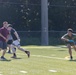 A group of young men are playing ultimate frisbee on a turf athletic field. One player on the right is holding a blue frisbee in a ready stance, while two players on the left appear to be defending or positioning themselves. The setting appears to be a school or recreational sports facility surrounded by trees.