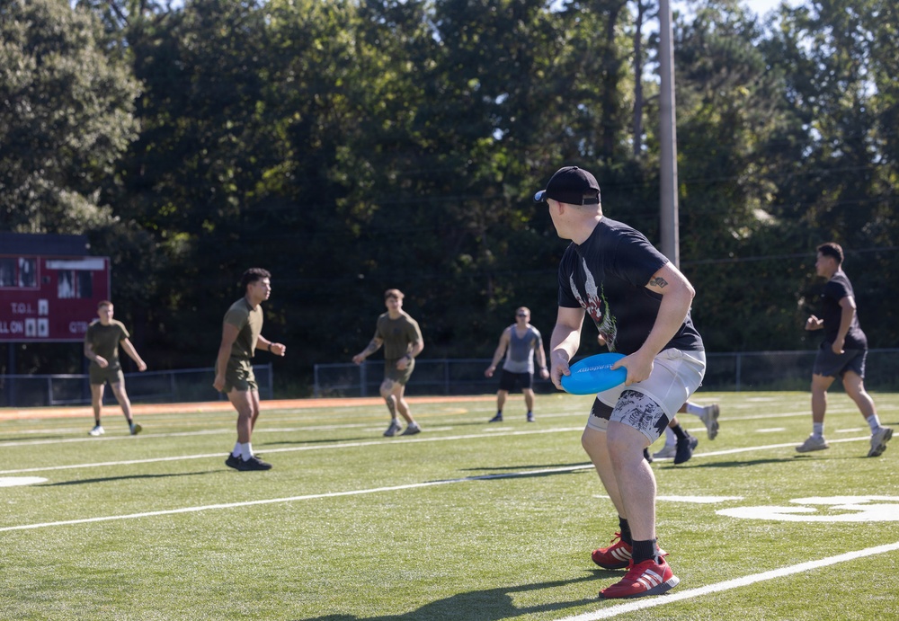A group of young men are playing Ultimate Frisbee on an artificial turf football field on a sunny day. The foreground player, wearing a black t-shirt, white shorts, and red Adidas shoes, is holding a blue frisbee in a ready stance. Several other players in olive/military-style clothing are visible in the background on the field.