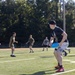 A group of young men are playing Ultimate Frisbee on an artificial turf football field on a sunny day. The foreground player, wearing a black t-shirt, white shorts, and red Adidas shoes, is holding a blue frisbee in a ready stance. Several other players in olive/military-style clothing are visible in the background on the field.
