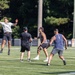 A group of men are playing ultimate frisbee on an outdoor turf field. One player is airborne, having just caught or thrown a white frisbee with arms outstretched. Several other players are positioned around him in athletic stances, suggesting active gameplay.