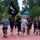 A group of military personnel and civilians are running together on a red outdoor track, with one participant carrying a POW/MIA flag. The event appears to be a commemorative run or memorial event honoring prisoners of war and those missing in action. The setting includes trees, a playground structure in the background, and overcast skies suggesting light rain.